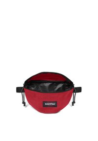 Punainen kangaspussi, jolla on pyöreä muoto, Musta sisävuori, säädettävä naru ja etuosassa brändilogo.