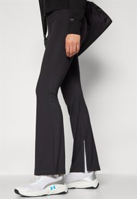Fekete, sportos leggings, melyek bővülő szabással rendelkeznek, oldalsó hasítékokkal a bokánál és kontrasztos varrással. Fehér és ezüst sportcipővel párosítva.