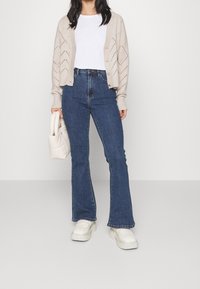 Nainen, joka on pukeutunut beigen neulotun neuletakin, valkoisen t-paidan, korkeavyötäröisiin sinisiin flare-farkkuihin ja valkoisiin plateausandaaleihin, pitäen kädessään pientä valkoista käsilaukkua.