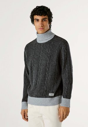 Pull en maille torsadée gris avec un col haut à côtes ; présente des poignets et un ourlet en gris clair contrastant, fabriqué en tissu de laine texturé.