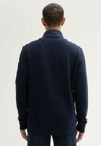 Pulover lung cu mâneci lungi de culoare bleumarin, cu guler înalt, textură ribbită și detalii cu buzunare laterale. Potrivire confortabilă, țesătură netedă.