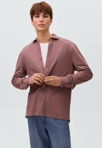 Giovane uomo che abbottona una camicia a maniche lunghe color rosa polveroso sopra una t-shirt bianca, indossando jeans blu, in piedi contro uno sfondo grigio chiaro uniforme.