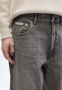 Γκρι τζιν από τζιν ύφασμα με άνετη γραμμή, με ορατό λογότυπο στον μπροστινό τσέπης και πλεκτή πουλόβερ σε κρεμ απόχρωση.