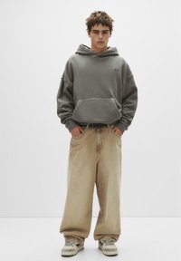 Grauer Kapuzenpullover mit Kängurutasche, kombiniert mit lockeren beigen Jeans und grauen Sneakers. Einfaches Design und lässige Passform.