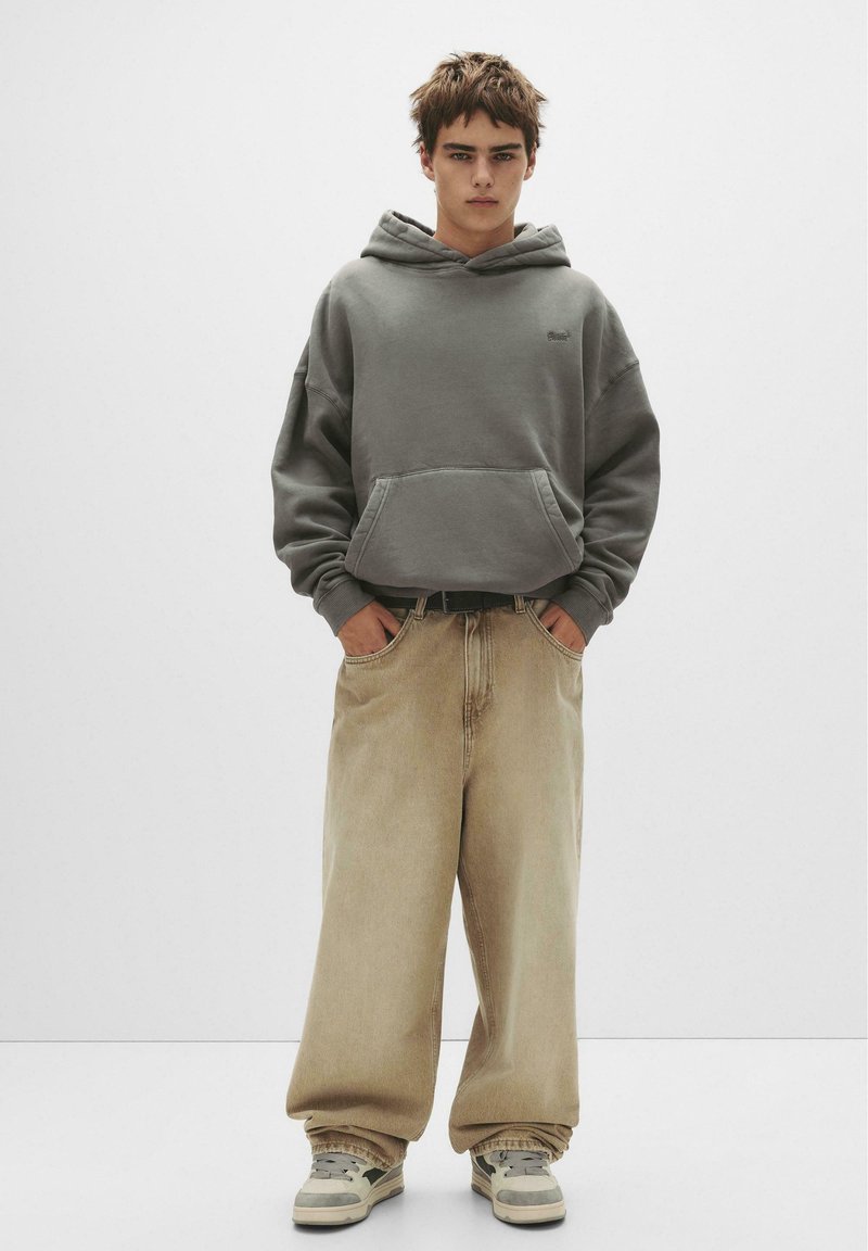 Grauer Kapuzenpullover mit Kängurutasche, kombiniert mit lockeren beigen Jeans und grauen Sneakers. Einfaches Design und lässige Passform.