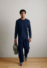 Tumši zila krekls ar garām piedurknēm, tumšas džinsa bikses un sporta apavi. Cilvēks turot zaļu somu, stāv uz koka grīdas neitrālas krāsas fonā.