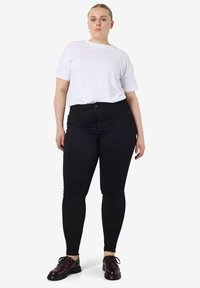 Mujer de piel clara y cabello rubio recogido, viste una camiseta blanca de manga corta, pantalones ajustados negros y zapatos oscuros de cordones, de pie.