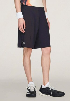 Persona che indossa pantaloncini neri Y-3, scarpe da ginnastica bianche e nere, polsini bianchi e una maglia colorata, in piedi davanti a uno sfondo semplice.