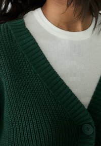 Green knitted cardigan with a V-neck and ribbed texture, featuring a button closure and worn over a white ribbed long-sleeve shirt.