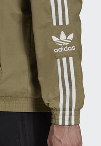 Olivgrön jacka med vita horisontella ränder på ärmarna, prydd med Adidas-logotypen broderad på vänster arm. Slät textur.