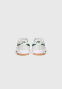Hvite Nike-sneakers med grønne detaljer og såle i gummi. De har en glatt læroverdel og borrelås-lukking.