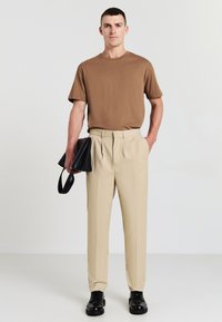 Uomo che indossa una t-shirt marrone e pantaloni beige, con una borsa in pelle nera in una mano, in piedi davanti a uno sfondo chiaro e uniforme.