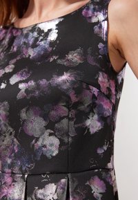 Czarna sukienka w kwiaty z metalicznym połyskiem, z fioletowymi i srebrnymi kwiatami. Materiał ma teksturowane wykończenie i usztywnioną konstrukcję.