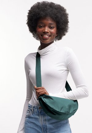 Lächelnde Frau mit natürlichem Haar trägt weißen Rollkragenpullover und Jeans, hält den grünen Riemen ihrer Umhängetasche über der Schulter vor weißem Hintergrund.