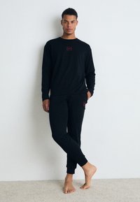 Ensemble de t-shirt à manches longues et de pantalon noirs en coton. Présente un petit logo rouge sur la poitrine et la poche, tous deux avec des poignets côtelés.