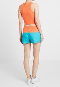 Oranžový bezrukávový crop top v kombinaci s tyrkysovými šortkami s elastickým pasem. Modelka má na sobě bílé tenisky.