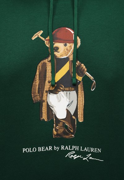 Plišasti medvedek oblečen v polo opremo s čelado, jakno, rokavicami in škornji, drži kladivo na temno zeleni tkanini z napisom »Polo Bear by Ralph Lauren«.