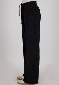 Zwarte, textuurvolle, wijde broek met een elastische tailleband, trekkoord, zijzakken en een ontspannen pasvorm. Ggedragen met lichtgekleurde sandalen.