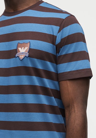 Άνδρας που φοράει κοντομάνικη μπλούζα με οριζόντιες ρίγες σε μπλε και καφέ χρώμα και ένα μικρό καφέ κεντητό έμβλημα με σχήμα ασπίδας του Emporio Armani Club στο στήθος.