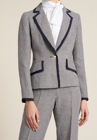 Blazer a cuadros personalizado en blanco y negro, con acentos en azul marino, botón dorado, dos bolsillos delanteros y solapas definidas.