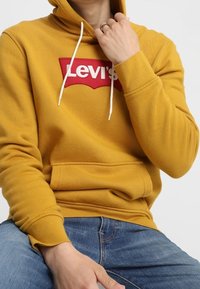 Felpa gialla senape con logo rosso Levi's sul petto, dotata di tasca a marsupio e lacci bianchi. Tessuto testurizzato, vestibilità casual.