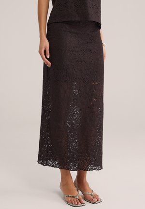 Femme portant une jupe midi en dentelle florale foncée et des sandales à talons motif peau de serpent, debout devant un fond beige uni.