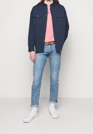 Uomo con camicia overshirt blu scuro abbottonata, t-shirt rosa, jeans chiari arrotolati, calze bianche e sneakers bianche in piedi davanti a uno sfondo semplice.