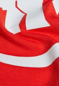 Röd t-shirt tyg med en slät textur; har en framträdande vit logotypdesign. Materialet verkar lätt och elastiskt.