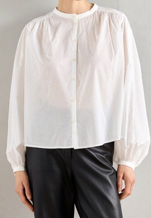 Blouse blanche boutonnée avec épaules froncées et longues manches bouffantes, associée à un pantalon en cuir noir sur une personne debout.