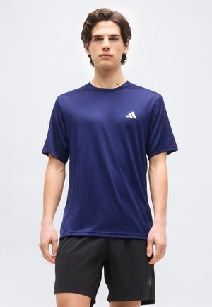 Junger Mann trägt ein navyblaues Adidas-T-Shirt und schwarze Shorts, steht vor einem schlichten hellen Hintergrund und blickt nach vorne.