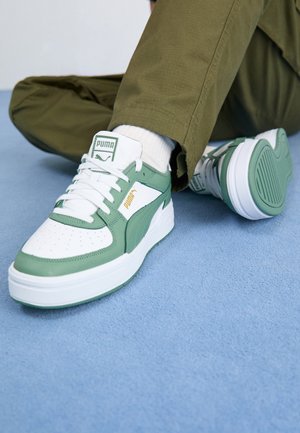 Osoba nosząca białe i zielone buty sportowe Puma z białymi skarpetkami i oliwkowo zielonymi spodniami, siedząca na jasnoniebieskiej powierzchni.