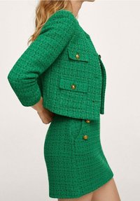 Blazer en tweed vert avec des boutons dorés et une jupe ajustée assortie. La texture est tissée avec un motif en grille, comportant des poches avant et un design court.