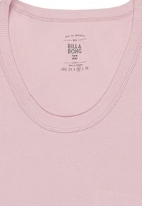 Top in cotone a coste di colore rosa chiaro, con scollatura tonda e etichetta che mostra "BILLA BONG", opzioni di taglia e istruzioni per la cura. Tessuto morbido.