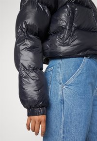 Manche de doudoune noire brillante avec poignet élastique, partiellement rentrée dans un jean en denim bleu taille haute avec poche arrière visible.