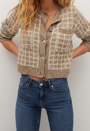 Mujer con un cárdigan de punto corto beige a cuadros con botones brillantes y jeans de mezclilla azul de talle alto, con las manos apoyadas en las caderas.