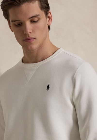 Mladi moški gleda navzdol, oblečen v belo majico z okroglim izrezom z majhnim črnim logotipom igralca polo na prsih.