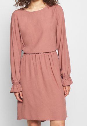 Femme portant une robe rose poudré à manches longues avec taille élastique et poignets à volants sur un fond clair uni.