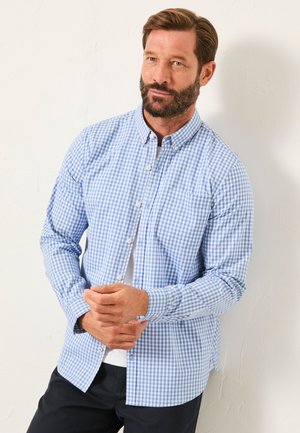 Man with beard wearing blue and white checkered button-up shirt over a white T-shirt, standing against a light wall.