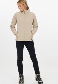 Beige softshell-jacka med hög krage och främre dragkedja, i kombination med svarta byxor och grå vandringskängor. Två sidofickor på jackan.