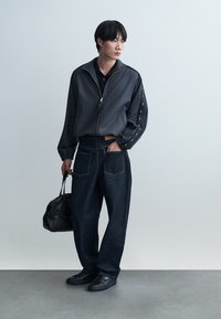 Tânăr purtând o jachetă închisă la culoare cu fermoar și blugi largi, ținând o geantă neagră de piele, stând în fața unui fundal simplu, deschis la culoare.