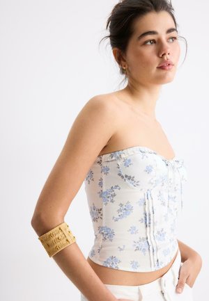 Jeune femme portant un corset fleuri blanc sans bretelles et un bracelet manchette beige texturé, regardant sur le côté.