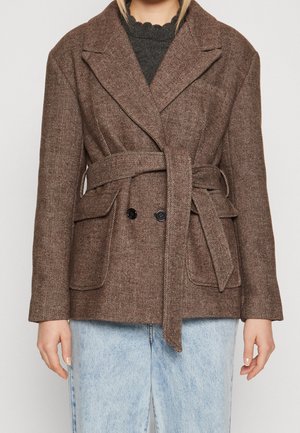 Woman wearing a brown herringbone belted wool blazer over a black scalloped-edge top and light blue jeans.