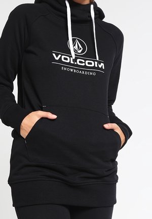 Person iført en sort Volcom snowboard-hoodie med hvide snore og hænder i forlommen.
