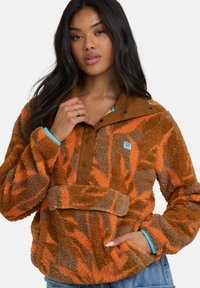 Vrouw in een bruin en oranje geblokt fleece jasje met blauwe bies, het kraag met één hand vasthoudend en de andere hand in de zak.