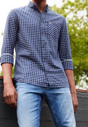 Homme portant une chemise à carreaux bleue avec les manches retroussées et un jean bleu clair, debout à l'extérieur sur un fond vert flou.