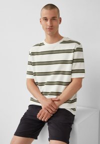 Gestreept t-shirt in wit met donkergroene horizontale lijnen, gemaakt van een gestructureerde stof, gecombineerd met donkere shorts, model staat met de armen gekruist.