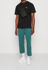 Černé tričko s malým logem, teal kordové kalhoty, bílé tenisky a černá crossbody taška. Neformální outfit s kontrastními texturami.