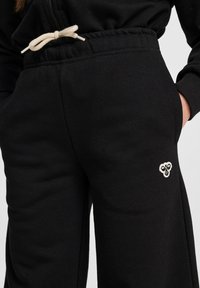Schwarze Jogginghosen mit elastischem Bund, cremefarbener Kordel und Seitentaschen. Mit kleinem, weißem, gesticktem Logo am Bein.