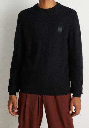 Homme portant un pull noir à manches longues avec un petit logo sur la poitrine et un pantalon marron ample, debout devant un fond uni.