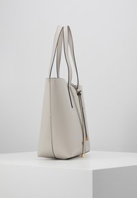 Lys beige læder tote taske med minimalistisk design, forlængede stropper og subtile hardware-accenter i guldtoner. Glat tekstur, vinklet form.